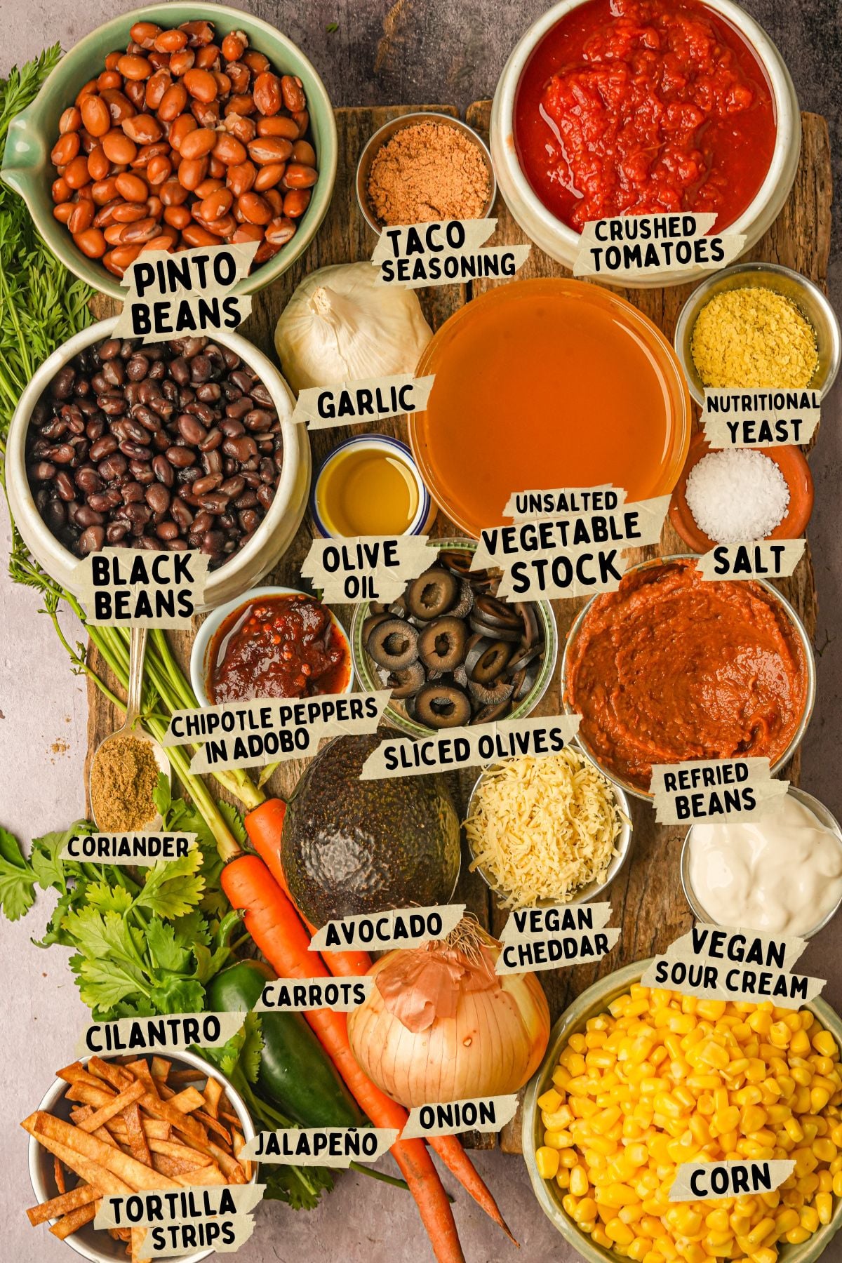 A variety of labeled ingredients for a vegan taco soup recipe are arranged on a table, including pinto and black beans, crushed tomatoes, taco seasoning, vegetable stock, nutritional yeast, spices, vegetables, vegan cheddar, and avocado.