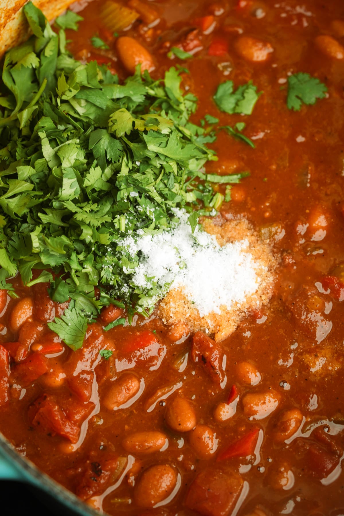 Pinto bean chili in a pot, topped with freshly chopped cilantro and piles of salt, with visible beans, tomatoes, and a rich, red sauce.