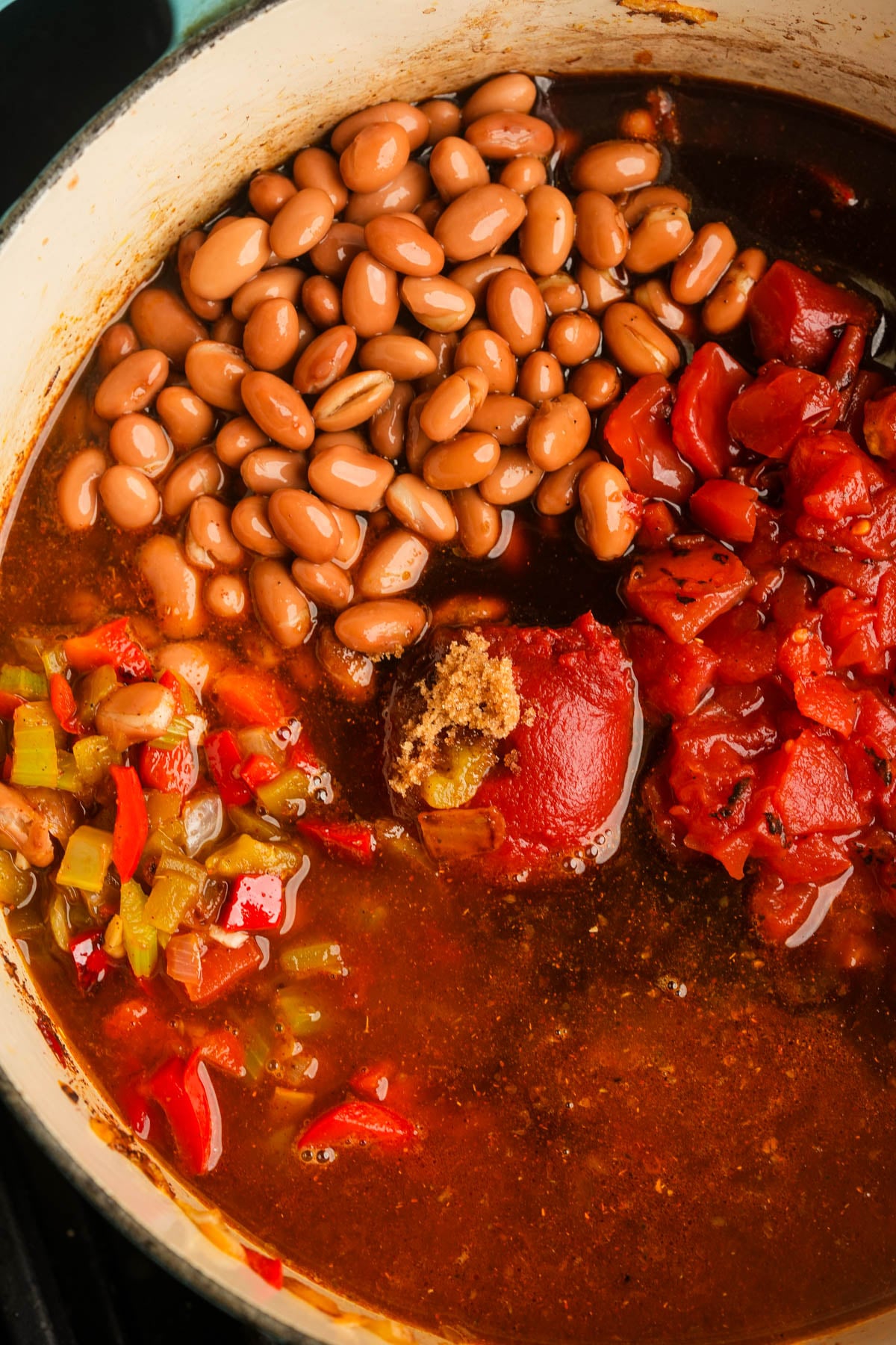 A close-up of a pot containing pinto beans, diced tomatoes, tomato paste, chopped bell peppers, and a rich broth, all ingredients separated and not yet stirred together.