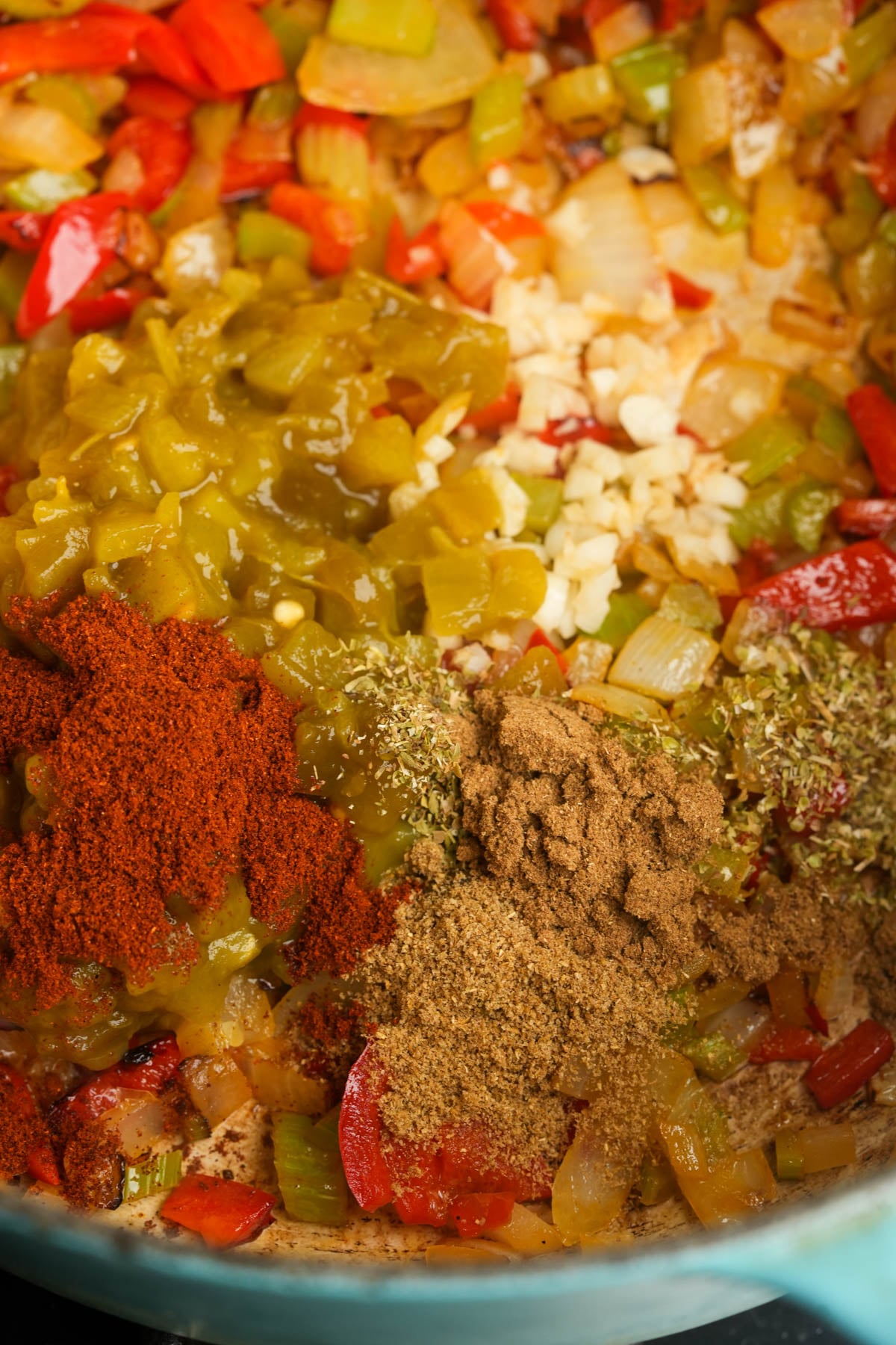 A close-up of chopped onions, celery, red bell peppers, green chilies, garlic, and assorted ground spices in a pot, ready to be mixed and cooked.
