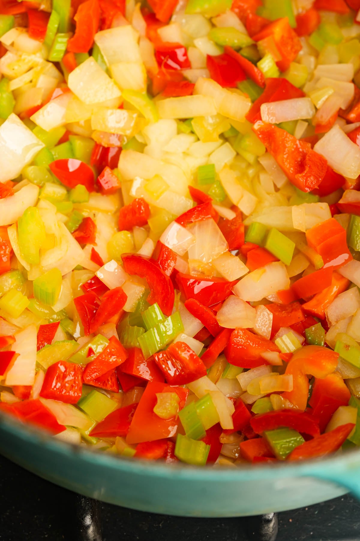 Chopped onions, red bell peppers, and celery sautéing in a pan, glistening with oil and starting to soften.