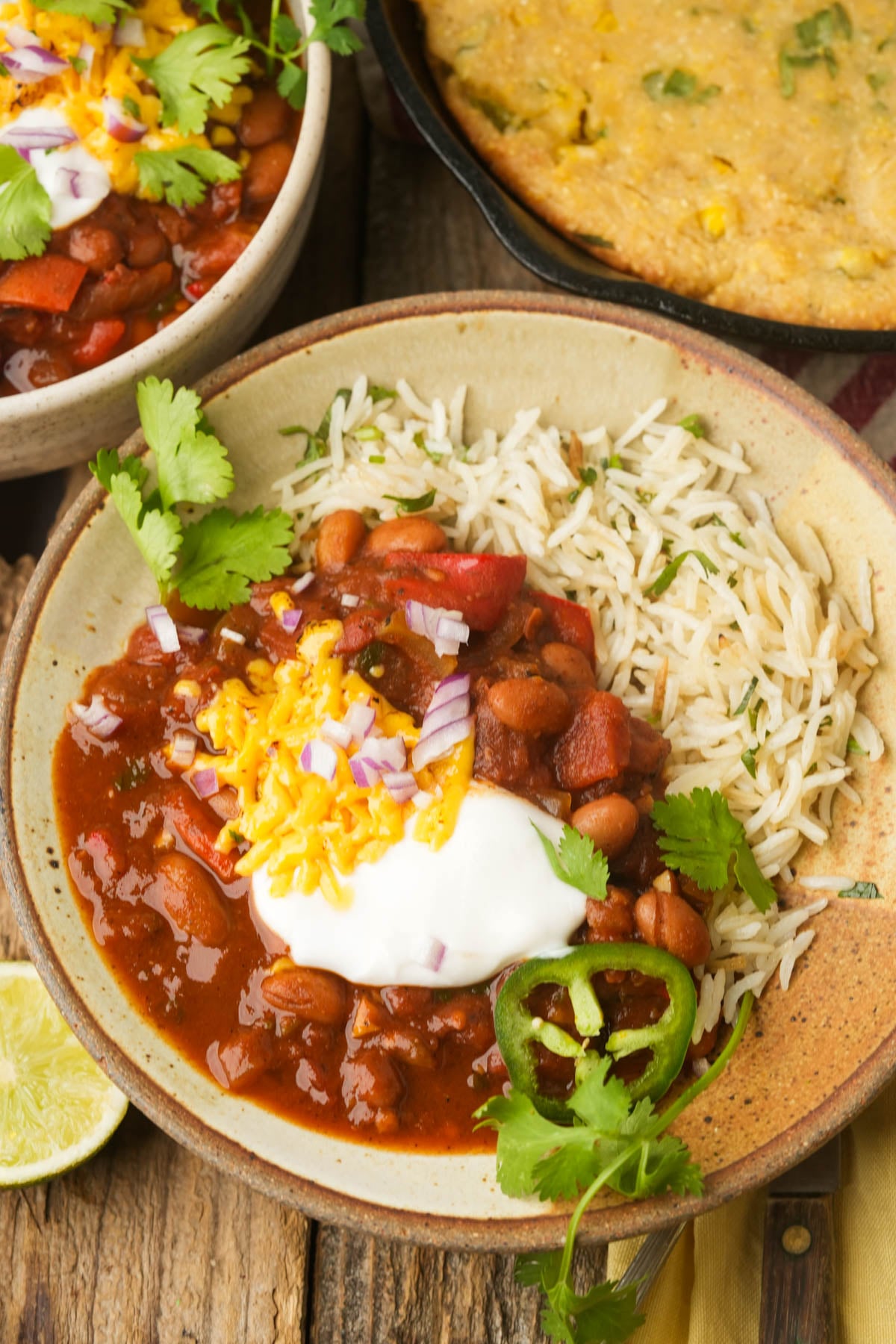 A bowl of pinto bean chili served with white rice, topped with shredded cheese, sour cream, chopped red onions, jalapeño slices, and cilantro. A lime wedge and a bowl of cornbread are nearby on a rustic wooden table.