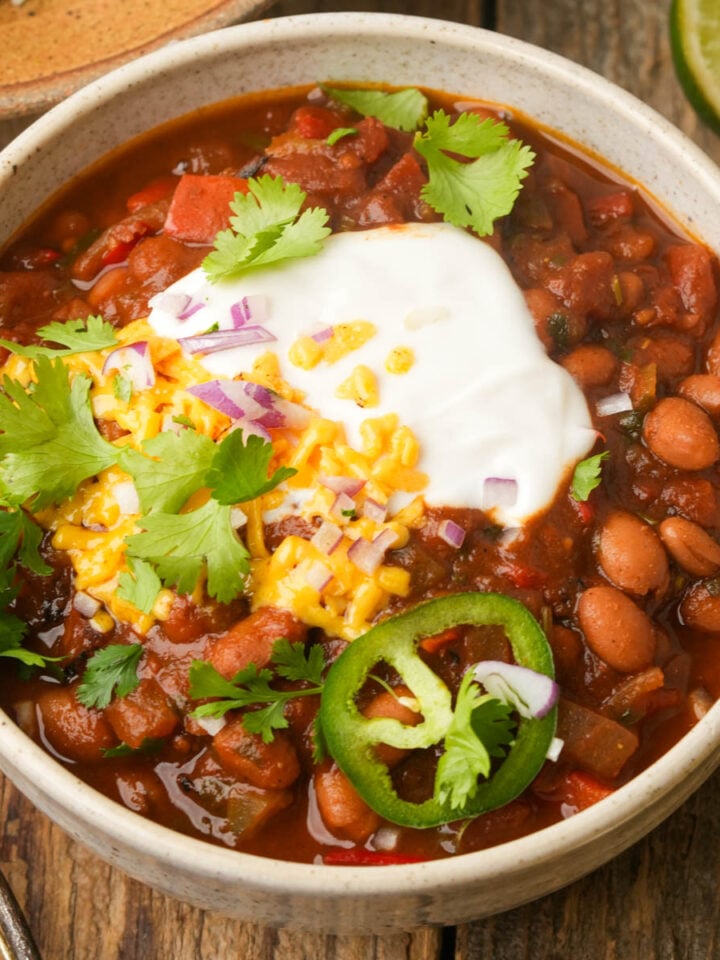 A bowl of pinto bean chili topped with sour cream, shredded cheese, chopped red onions, fresh cilantro, and sliced jalapeño, placed on a wooden table with a lime wedge nearby.