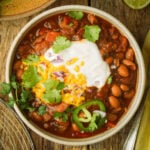 A bowl of pinto bean chili topped with shredded cheese, sour cream, chopped red onions, jalapeño slices, and fresh cilantro, sitting on a wooden table.