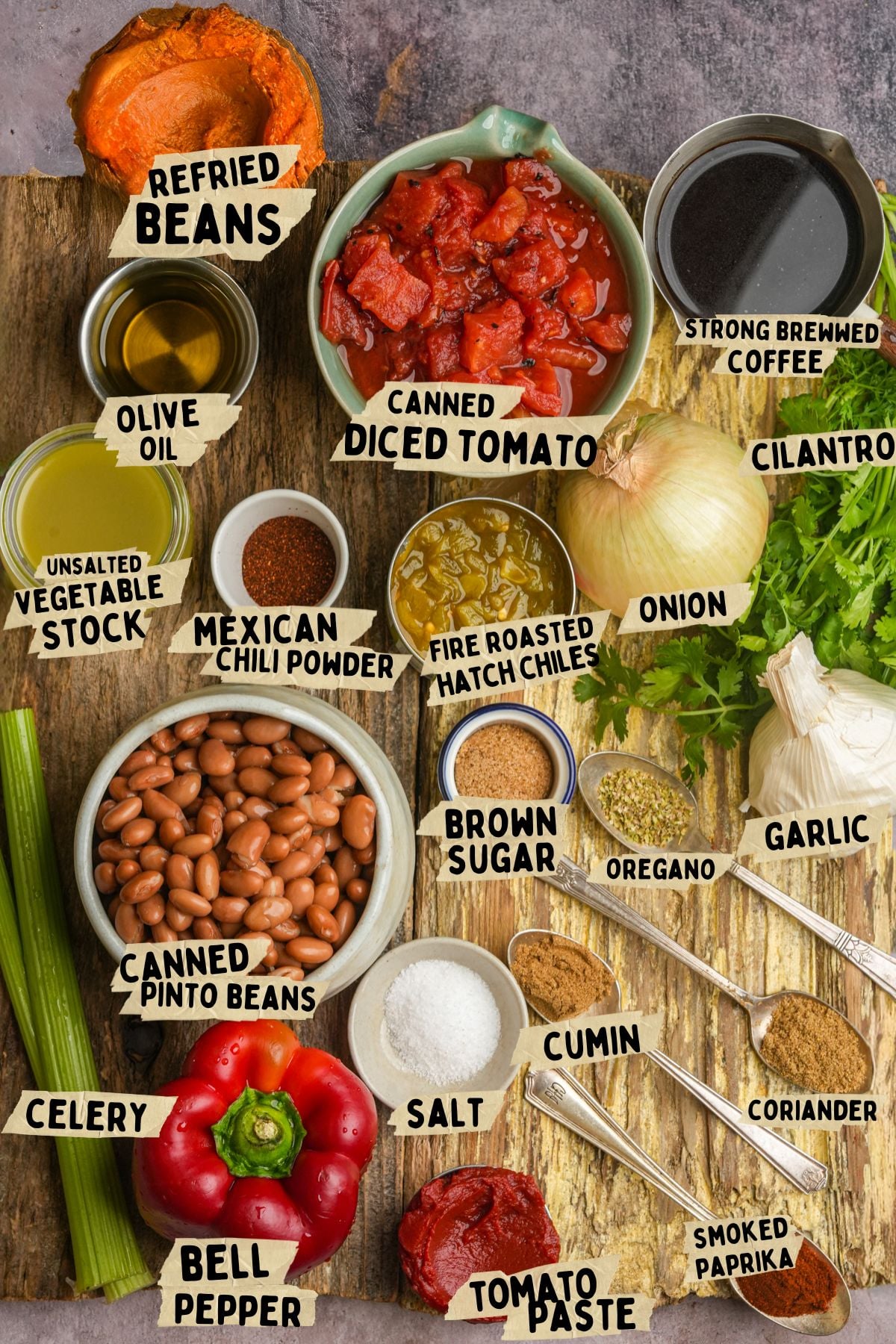 A variety of chili ingredients labeled on a wooden surface for a pinto bean chili recipe including refried beans, canned diced tomato, coffee, cilantro, olive oil, vegetable stock, chili powder, hatch chiles, onion, pinto beans, celery, bell pepper, spices, salt, and tomato paste.