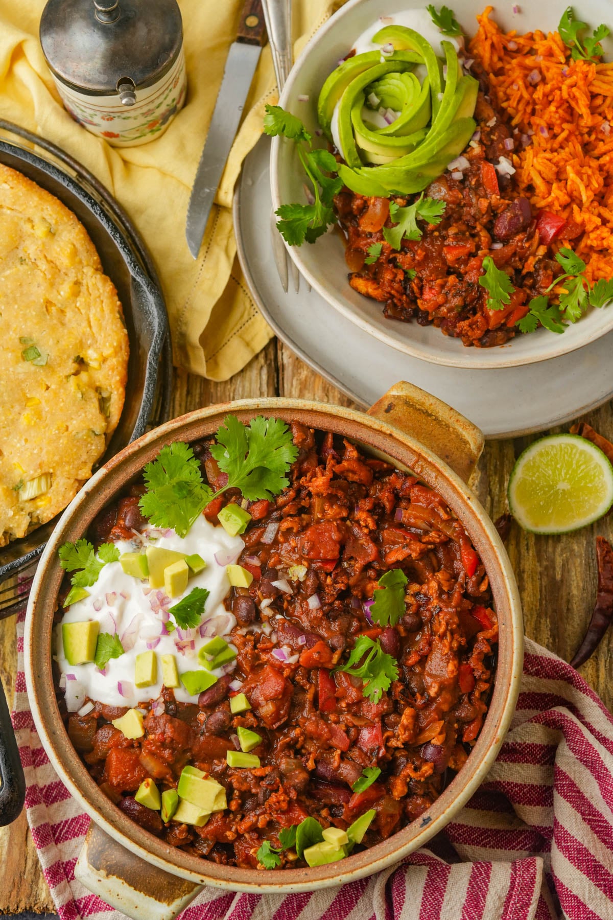 A bowl of tofu chili topped with cilantro, diced avocado, and sour cream sits on a wooden table next to cornbread, a plate of chili with rice, and half a lime. A striped napkin and pepper shaker are also visible.