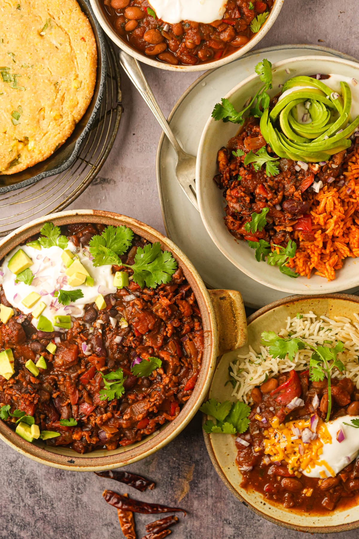 Assorted vegan chilies with toppings like avocado, sour cream, cilantro, and shredded cheese, served alongside cornbread, rice, and beans on a gray surface.