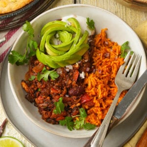 A bowl of tofu chili with beans and vegetables, served with orange rice and topped with sliced avocado shaped like a rose, fresh cilantro, and diced onions. A fork rests on the side of the bowl.