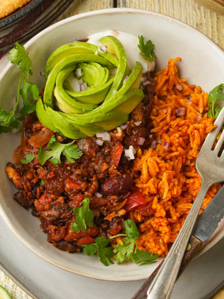 A bowl of tofu chili with beans and vegetables, served with orange rice and topped with sliced avocado shaped like a rose, fresh cilantro, and diced onions. A fork rests on the side of the bowl.