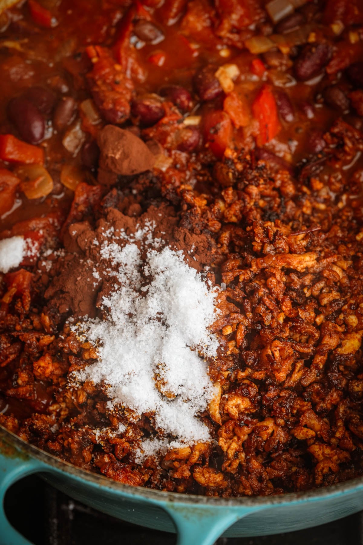 Ground tofu, red kidney beans, chopped tomatoes, and onions cooking in a pot, topped with cocoa powder and a mound of salt, being prepared to make tofu chili.