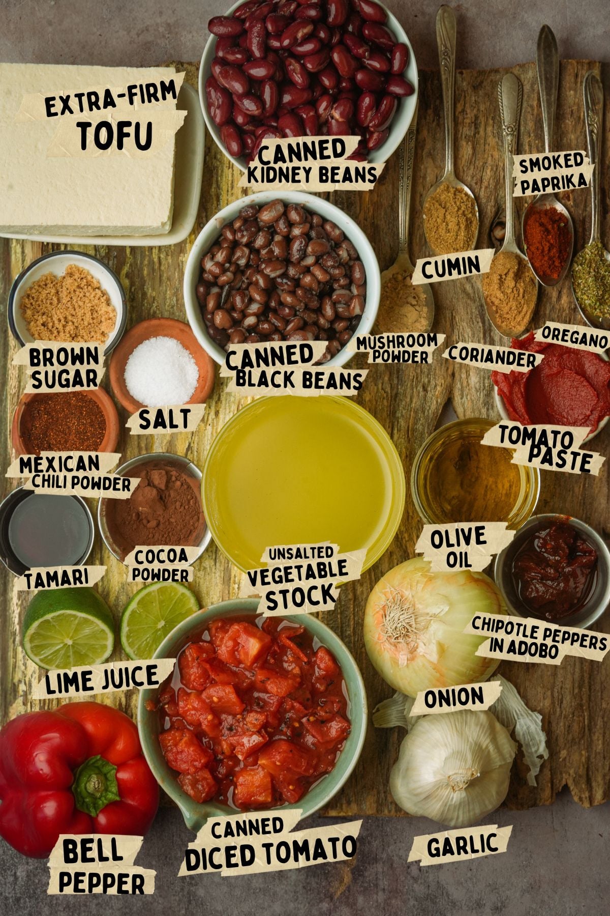 A variety of tofu chili ingredients arranged on a wooden surface, including tofu, bell pepper, spices, canned beans, diced tomato, vegetable stock, olive oil, onion, garlic, and labeled seasonings in bowls and spoons.