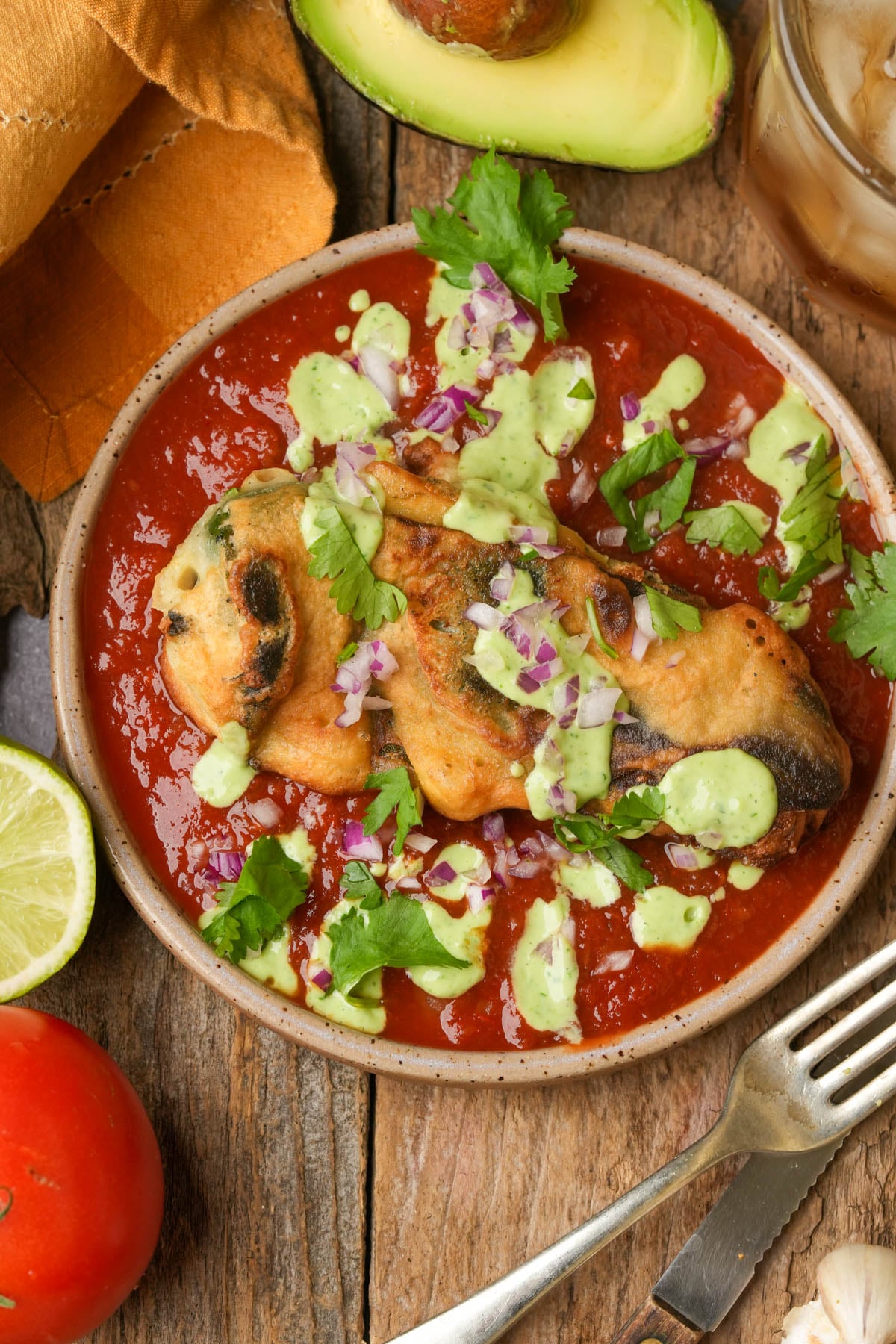 A plate of vegan chile relleno topped with green sauce, chopped red onion, and cilantro sits on red salsa. Surrounding the dish are a lime wedge, a tomato, half an avocado, a fork, and a glass of iced drink.