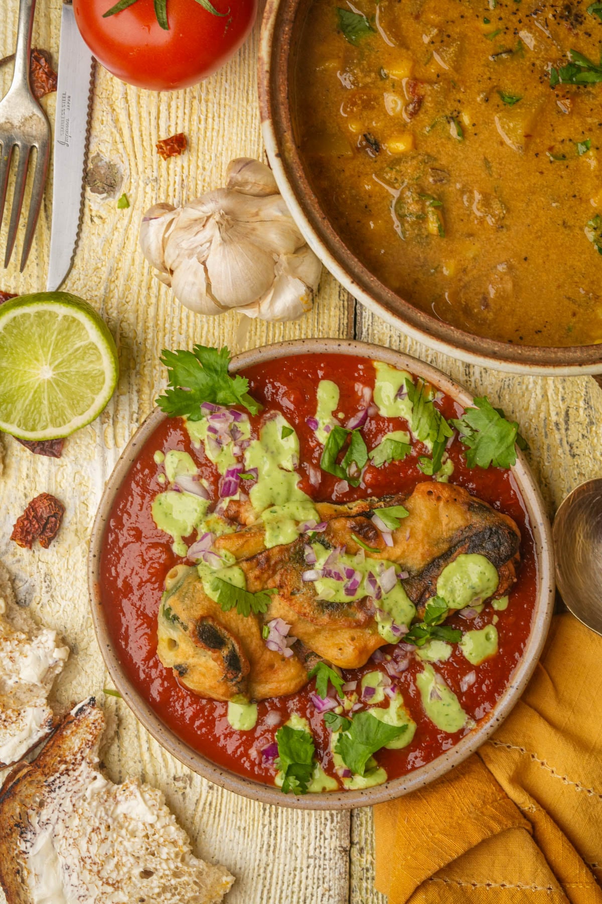 A plate of vegan chiles rellenos with red sauce, green sauce, and chopped onions, next to a bowl of vegan corn chowder, a lime, a tomato, garlic, and rustic bread on a wooden table.