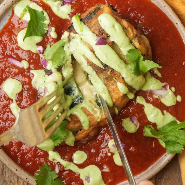 Vegan chiles rellenos topped with green sauce and cilantro sits in a bowl of red tomato sauce. A fork and knife are cutting into the pepper, revealing melted cheese and pieces of red onion.