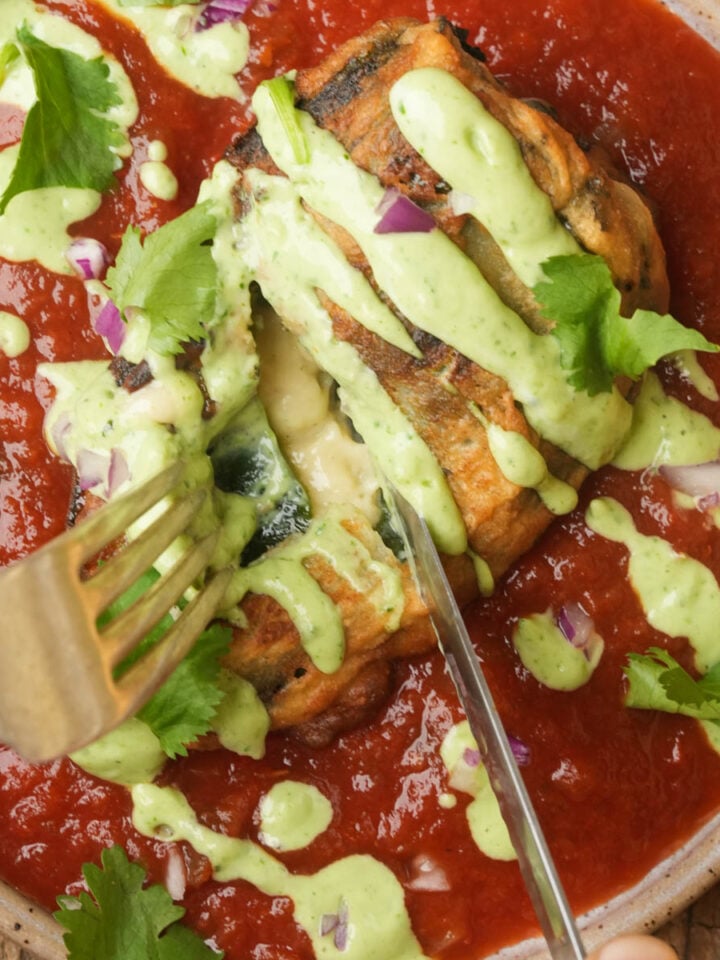 Vegan chiles rellenos topped with green sauce and cilantro sits in a bowl of red tomato sauce. A fork and knife are cutting into the pepper, revealing melted cheese and pieces of red onion.