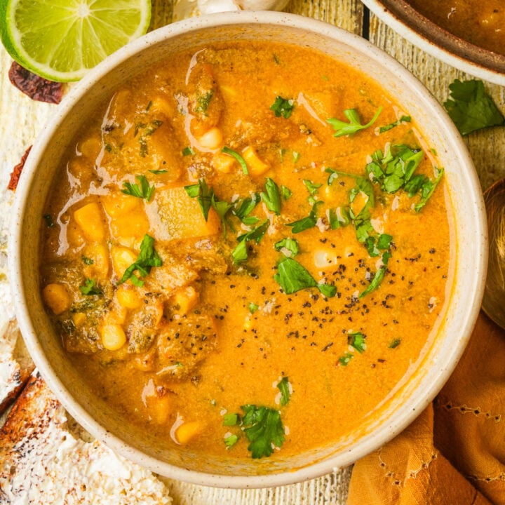 A bowl of vegan corn chowder garnished with chopped cilantro and black pepper, containing visible chunks of potatoes and corn, with a slice of lime and toasted bread on the side.