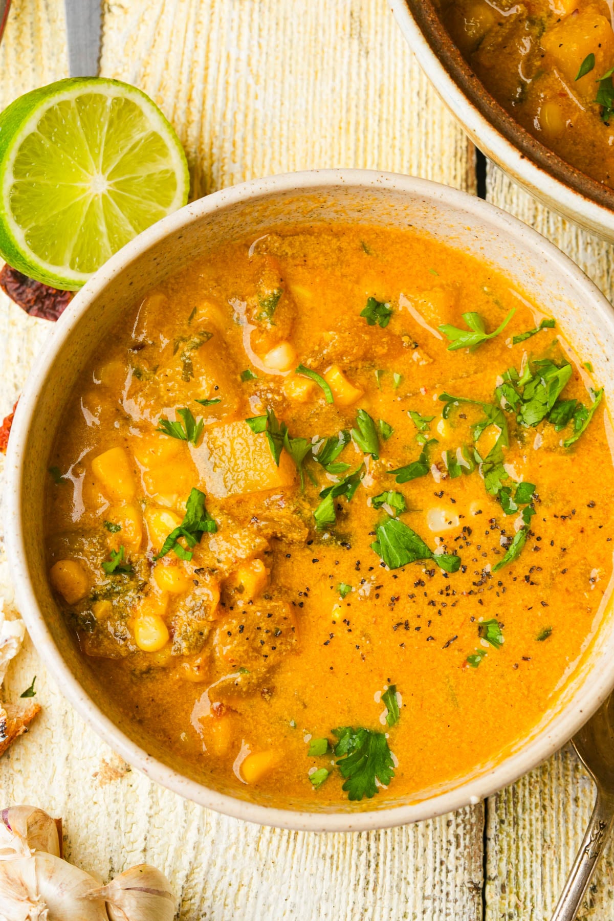 A bowl of creamy vegan corn chowder garnished with chopped cilantro and black pepper sits on a rustic wooden surface beside a halved lime and fresh garlic cloves.