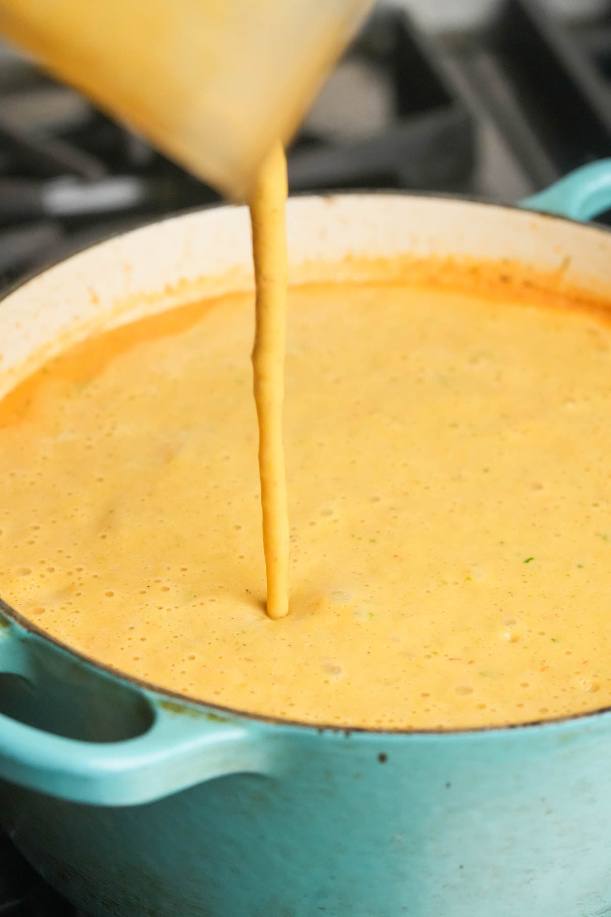A thick, creamy pureed portion of soup being poured into a large turquoise Dutch oven on a stovetop containing unblended vegan corn chowder.