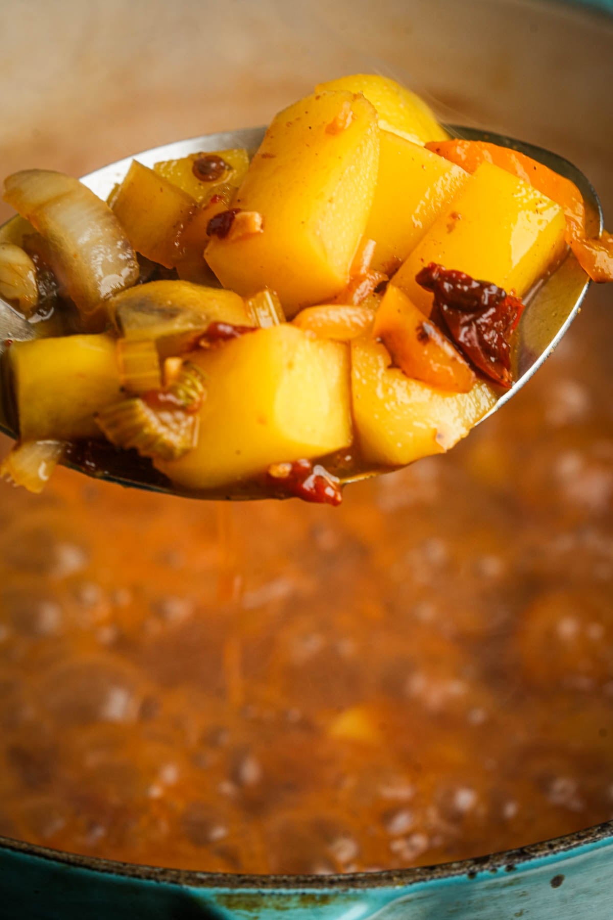 A close-up of a ladle holding chunky vegetable soup with potatoes, onions, and bits of chipotle above a pot of simmering vegan corn chowder.