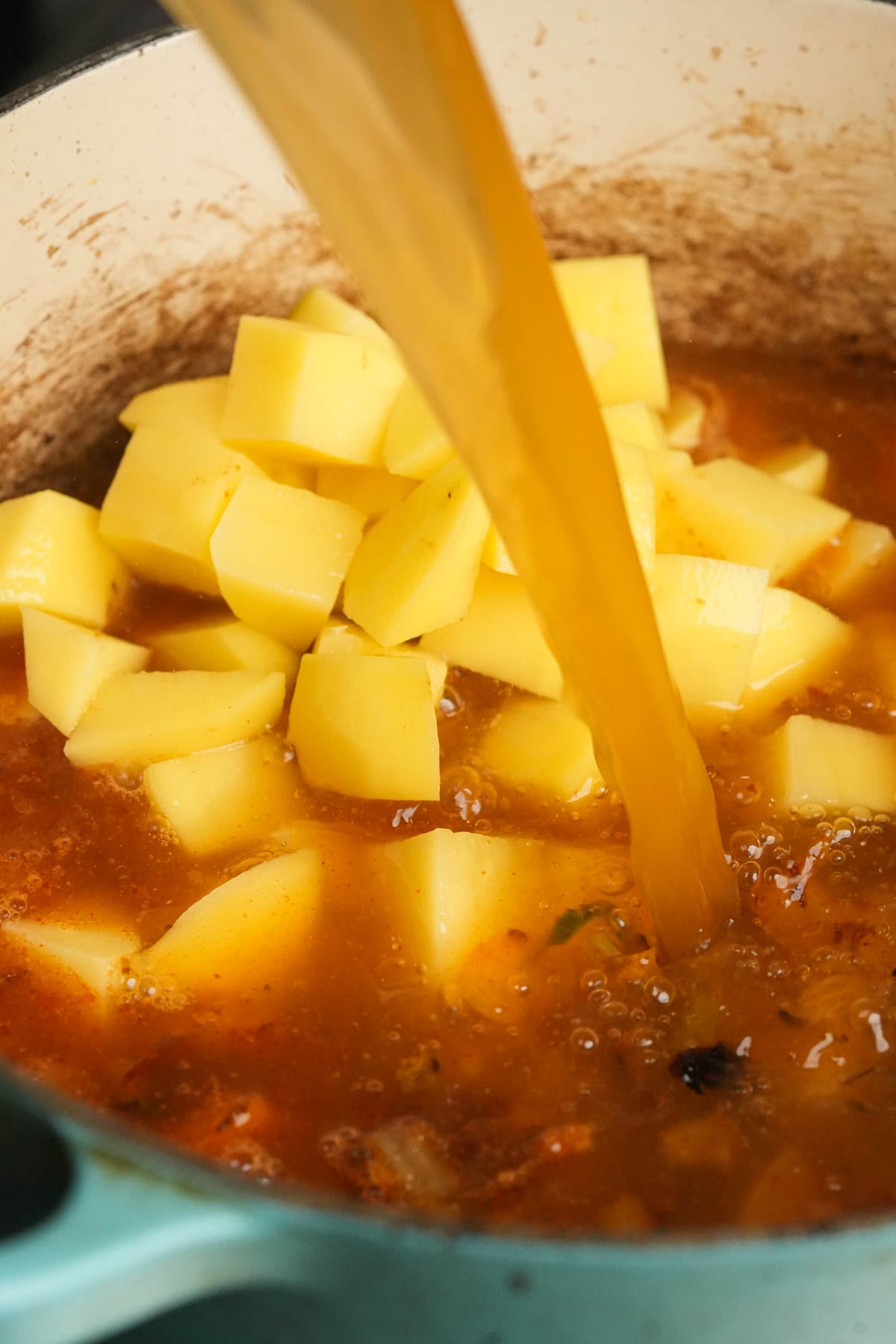 A stream of broth is being poured over diced potatoes in a pot filled with a rich, orange-brown liquid, making the base for vegan corn chowder.