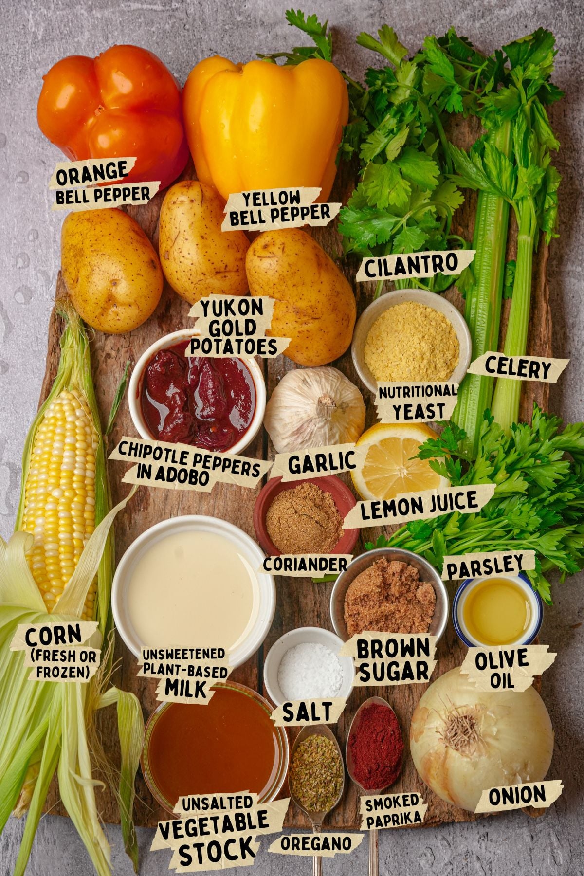 Assorted fresh and packaged ingredients for a vegan corn chowder recipe including orange and yellow bell peppers, Yukon gold potatoes, cilantro, celery, corn, chipotle peppers, garlic, nutritional yeast, plant-based milk, and spices.
