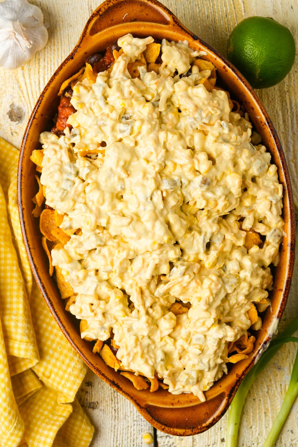 A brown oval casserole dish filled with a layered cheesy, creamy mixture over corn chips, sitting on a light wooden surface. Nearby are a lime, a yellow checkered napkin, green onions, and a garlic bulb.