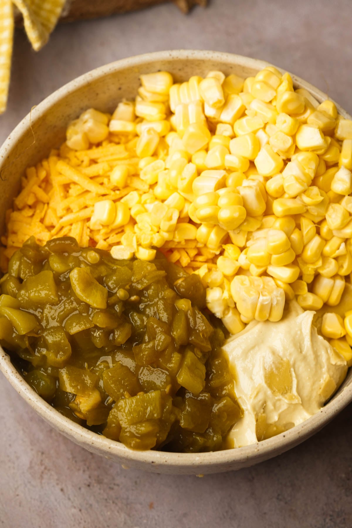 A bowl containing shredded vegan cheddar cheese, corn kernels, chopped green chilies, and a dollop of mayonnaise, arranged in separate sections on a neutral surface.