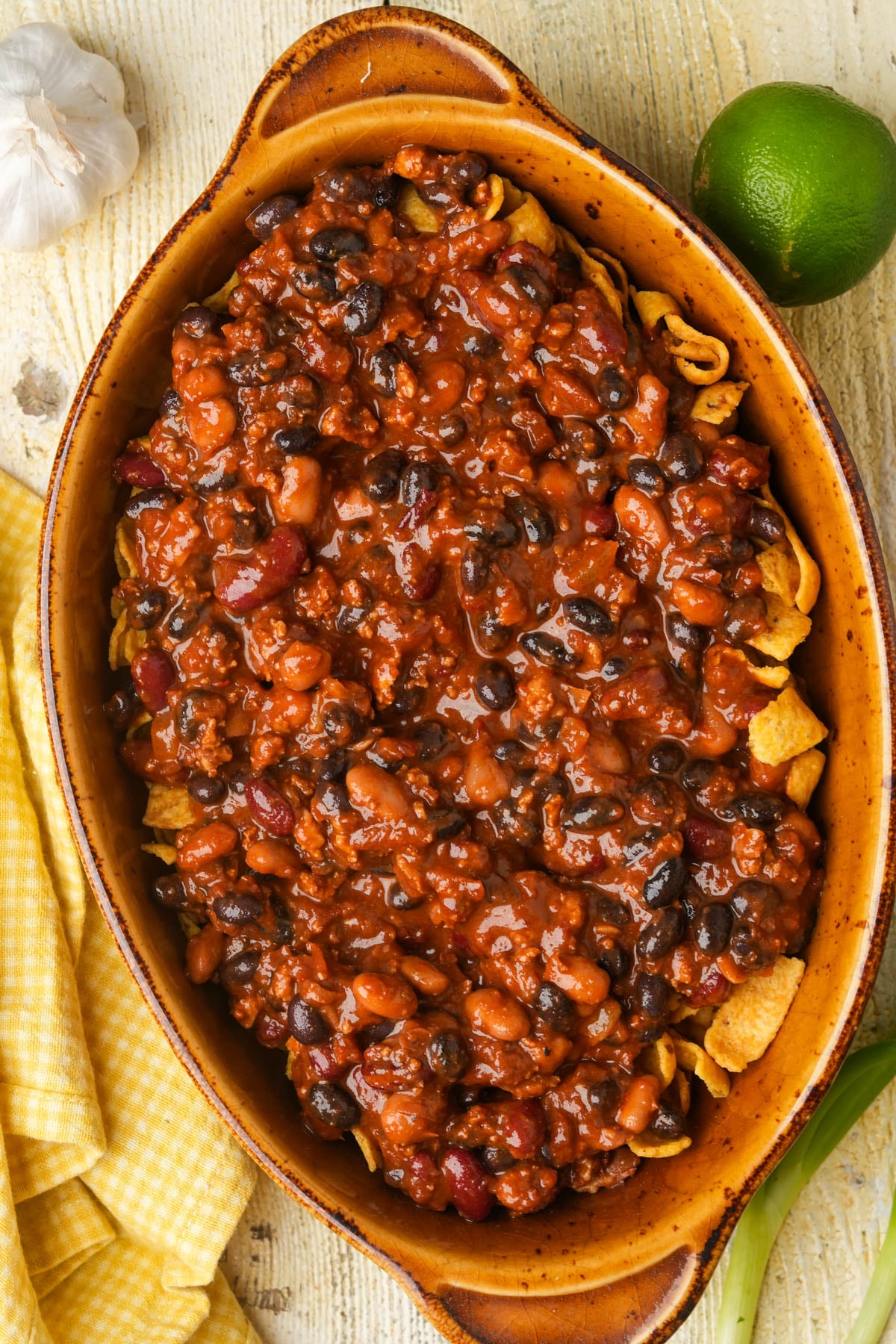 A baked casserole dish filled with chili made of beans and ground seitan, served over corn chips. The dish sits on a light wood surface with a lime, garlic, green onion, and a yellow checkered towel nearby.