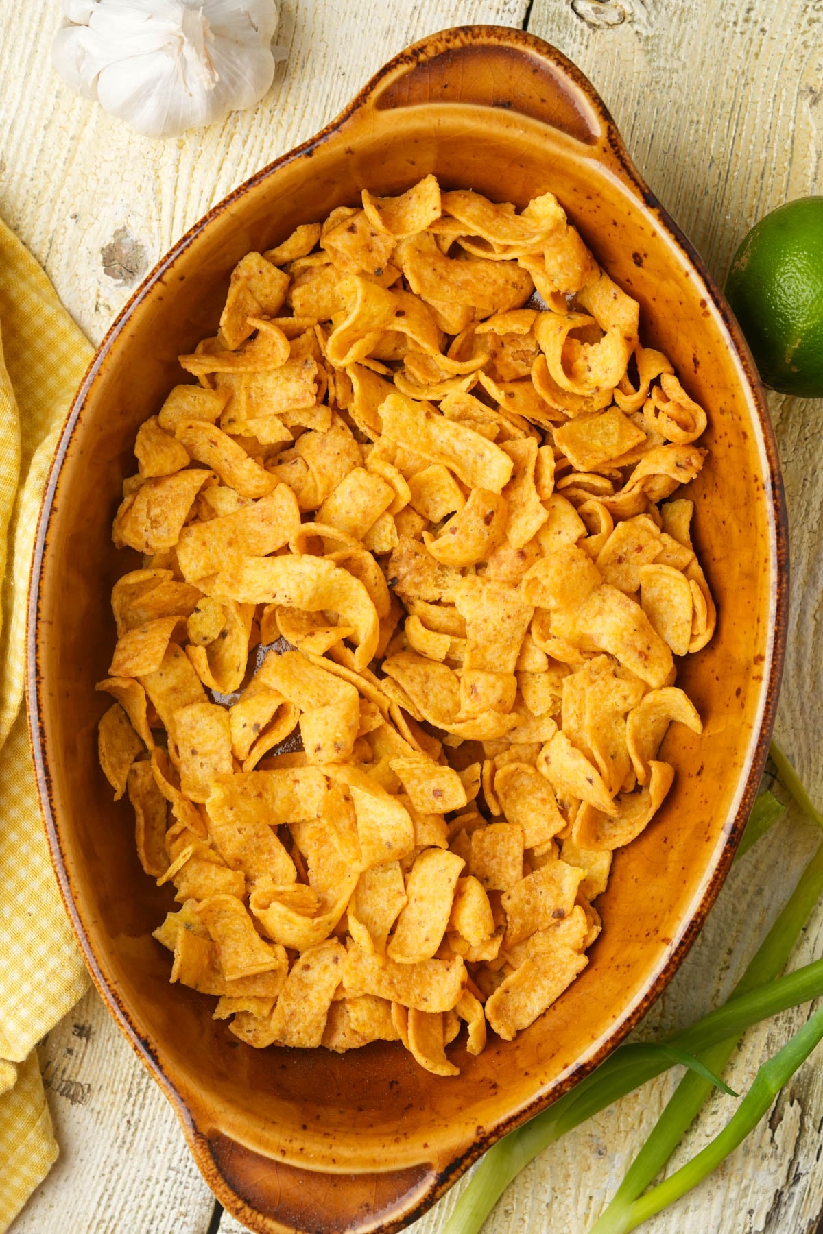 A brown oval dish filled with golden, seasoned corn chips sits on a rustic wooden surface, with a lime, garlic, green onions, and a yellow cloth nearby.