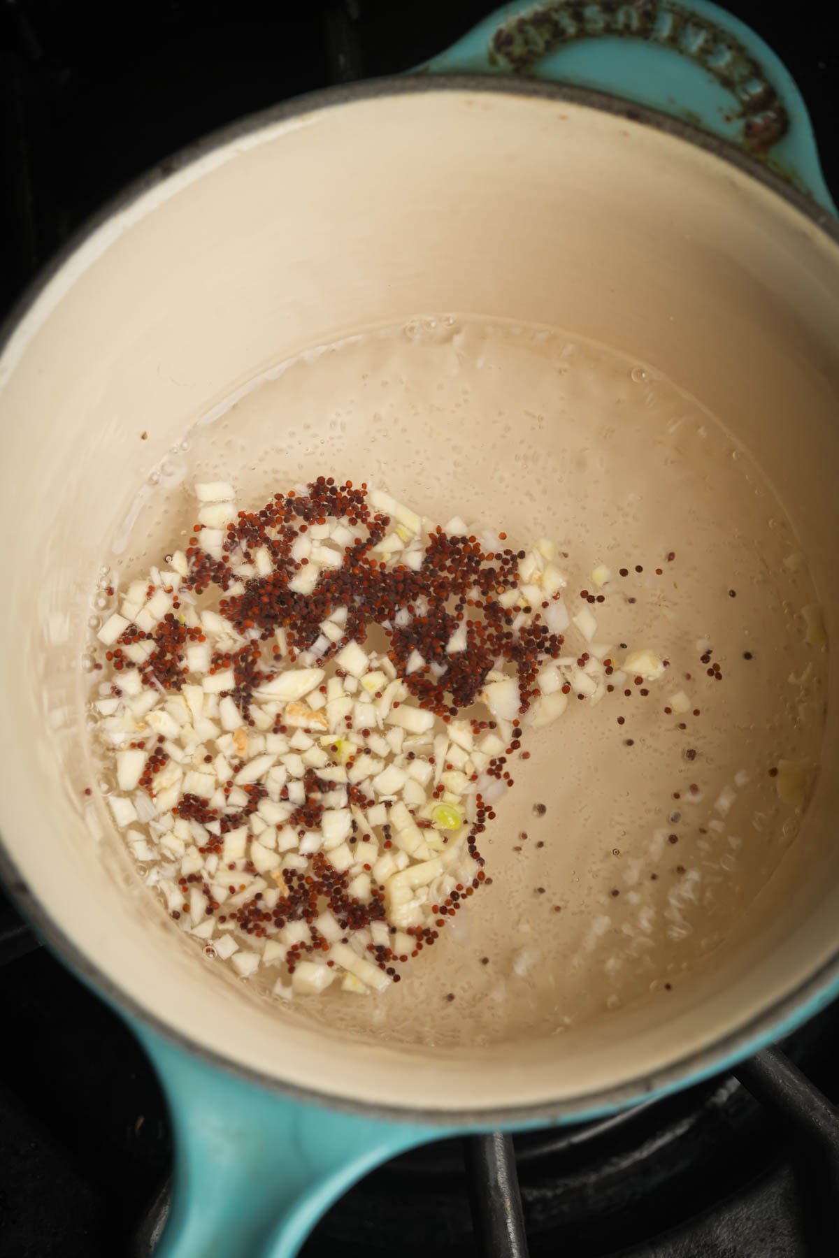 Chopped garlic and mustard seeds simmering in water inside a light-colored pot with a turquoise handle, on a stovetop-perfect aromatics for starting your Vegan Frito Pie Casserole.