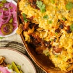 A cheesy vegan Frito pie casserole with beans, chips, and cilantro in a brown dish, next to a small bowl of pickled red onions and jalapeños, and a plate with avocado and sour cream.