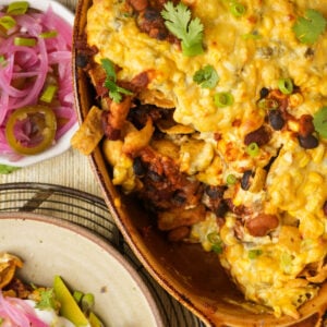 A cheesy vegan Frito pie casserole with beans, chips, and cilantro in a brown dish, next to a small bowl of pickled red onions and jalapeños, and a plate with avocado and sour cream.