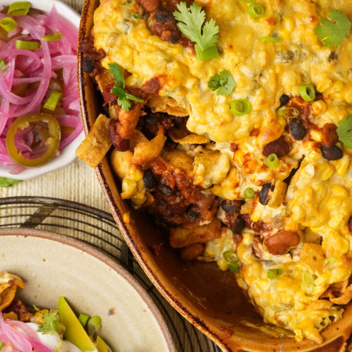A cheesy vegan Frito pie casserole with beans, chips, and cilantro in a brown dish, next to a small bowl of pickled red onions and jalapeños, and a plate with avocado and sour cream.