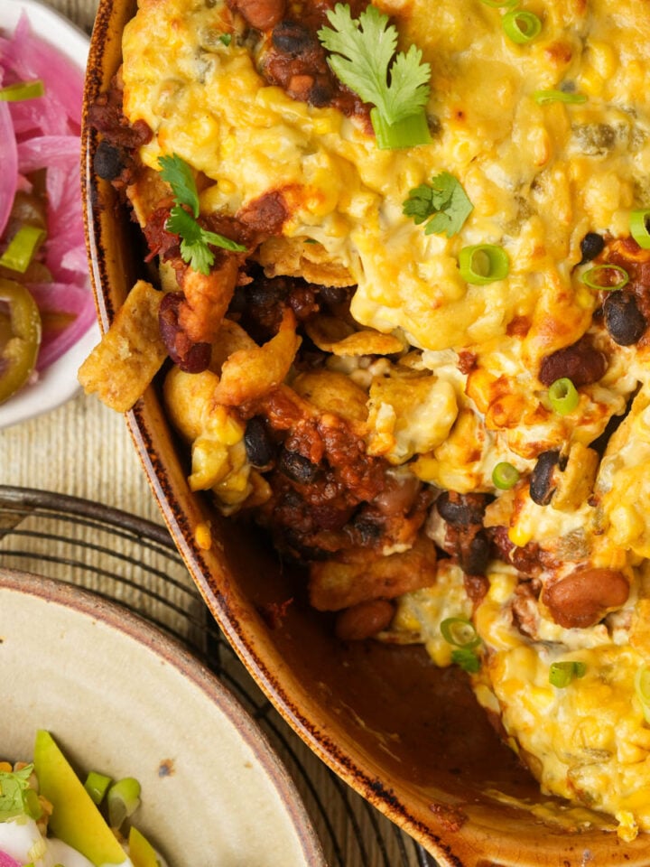 A cheesy vegan Frito pie casserole with beans, chips, and cilantro in a brown dish, next to a small bowl of pickled red onions and jalapeños, and a plate with avocado and sour cream.