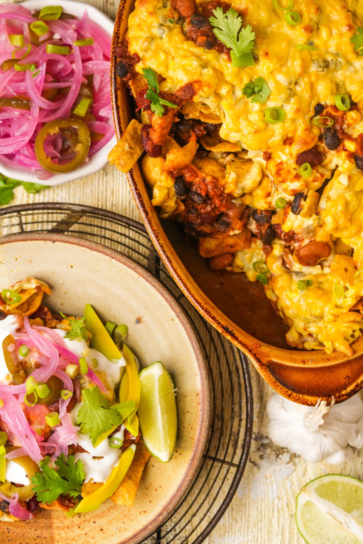 A colorful spread with a baked cheesy vegan Frito pie, served beside a plate of nachos topped with avocado, pickled onions, sour cream, and lime wedges. Fresh herbs and jalapeños garnish the dishes.