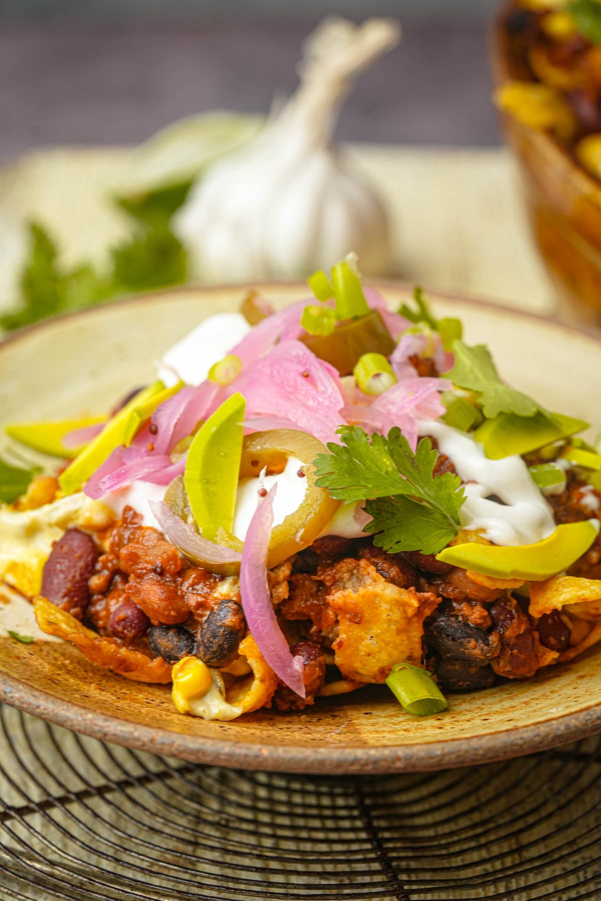 A plate of vegan Frito pie topped with chili, black beans, corn, pickled onions, avocado slices, jalapeños, sour cream, cilantro, and sliced green onions, with a garlic bulb and herbs in the background.