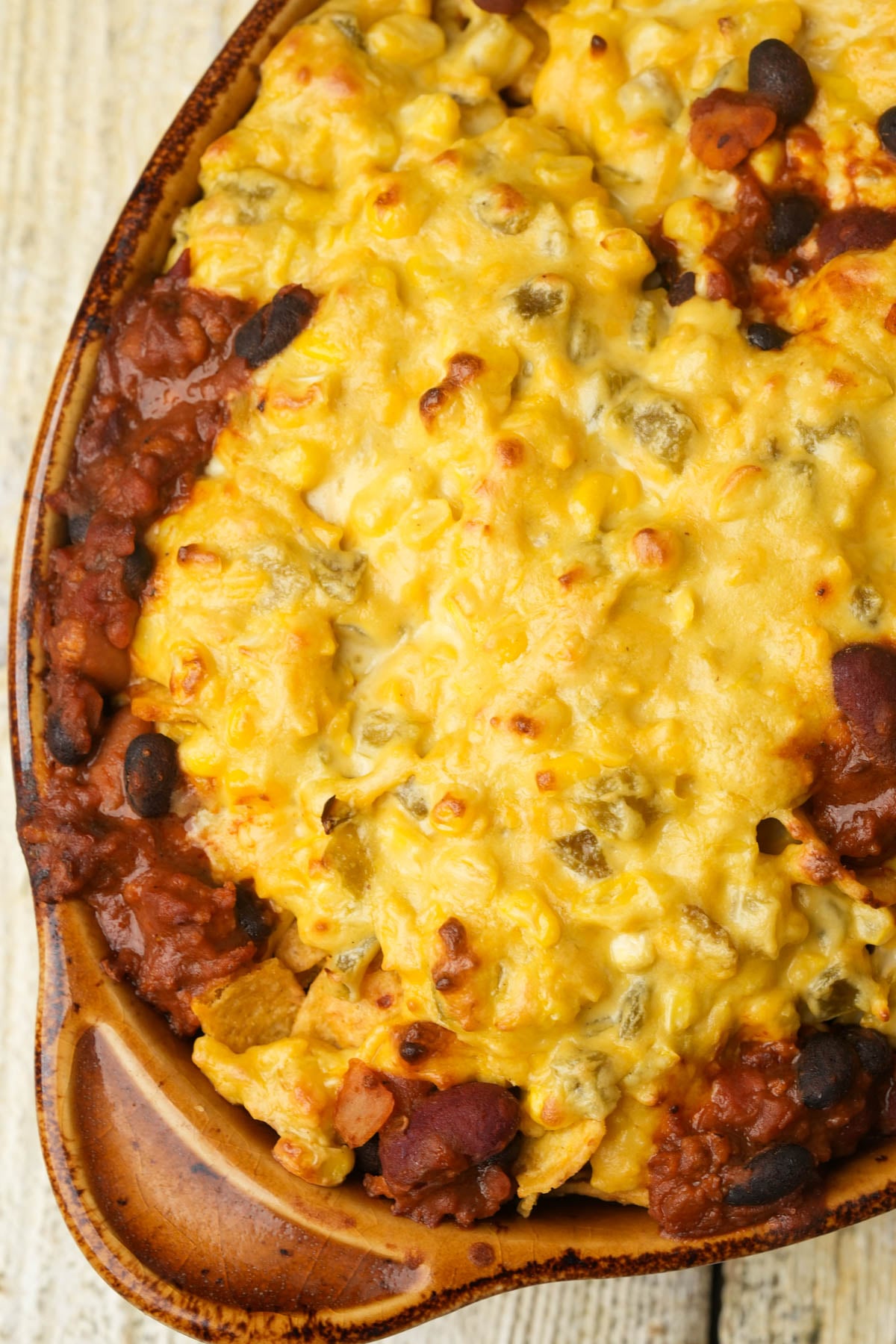 Close-up of a baked vegan Frito pie with melted cheese on top. The casserole appears to have a mix of beans, corn, and possibly ground meat beneath the golden, bubbly cheese layer.