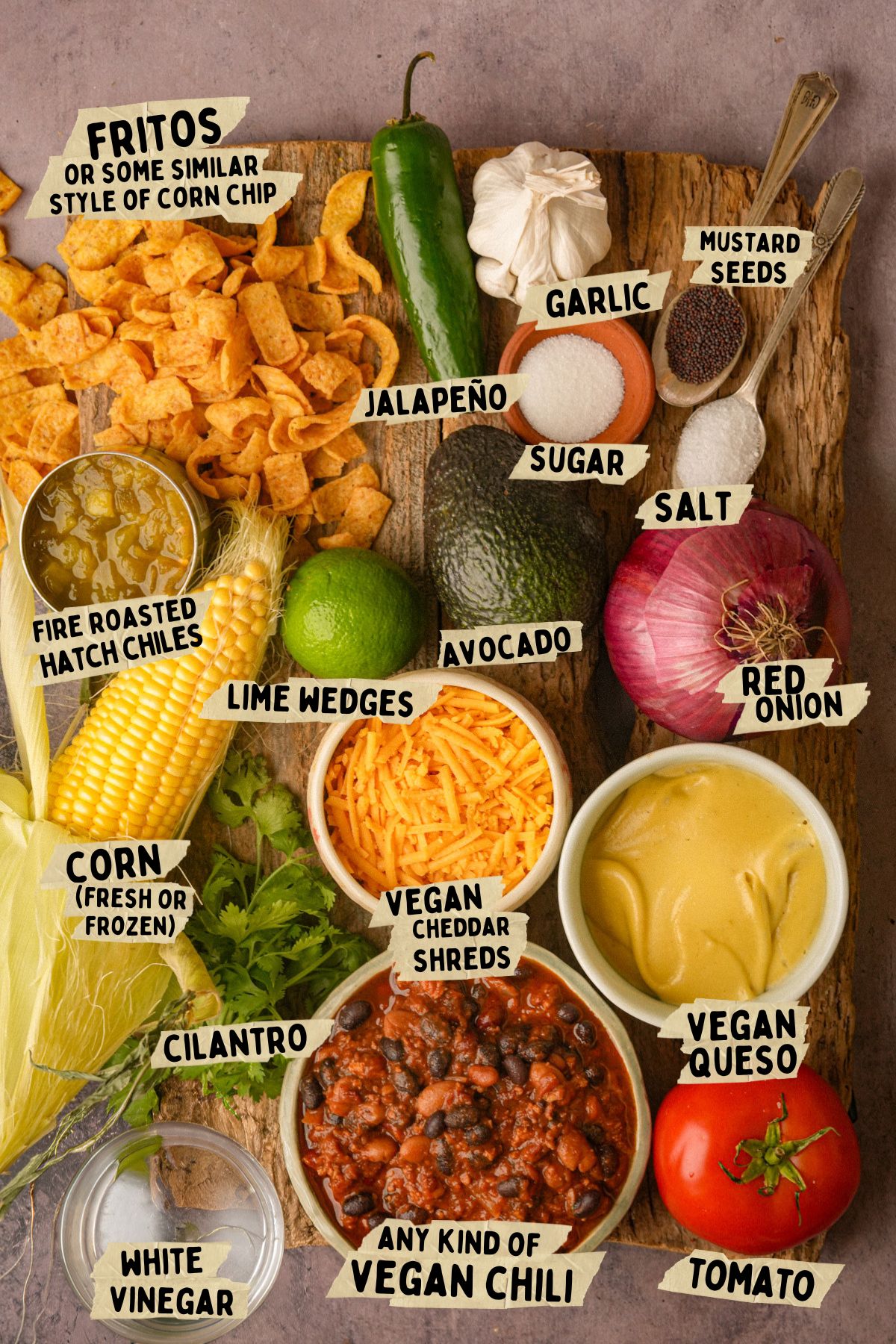 A top-down view of ingredients on a wooden surface for a vegan Frito pie recipe including Fritos, chili, vegan queso, corn, avocado, red onion, lime wedges, cilantro, jalapeño, garlic, sugar, salt, mustard seeds, tomato, and fire-roasted Hatch chiles.