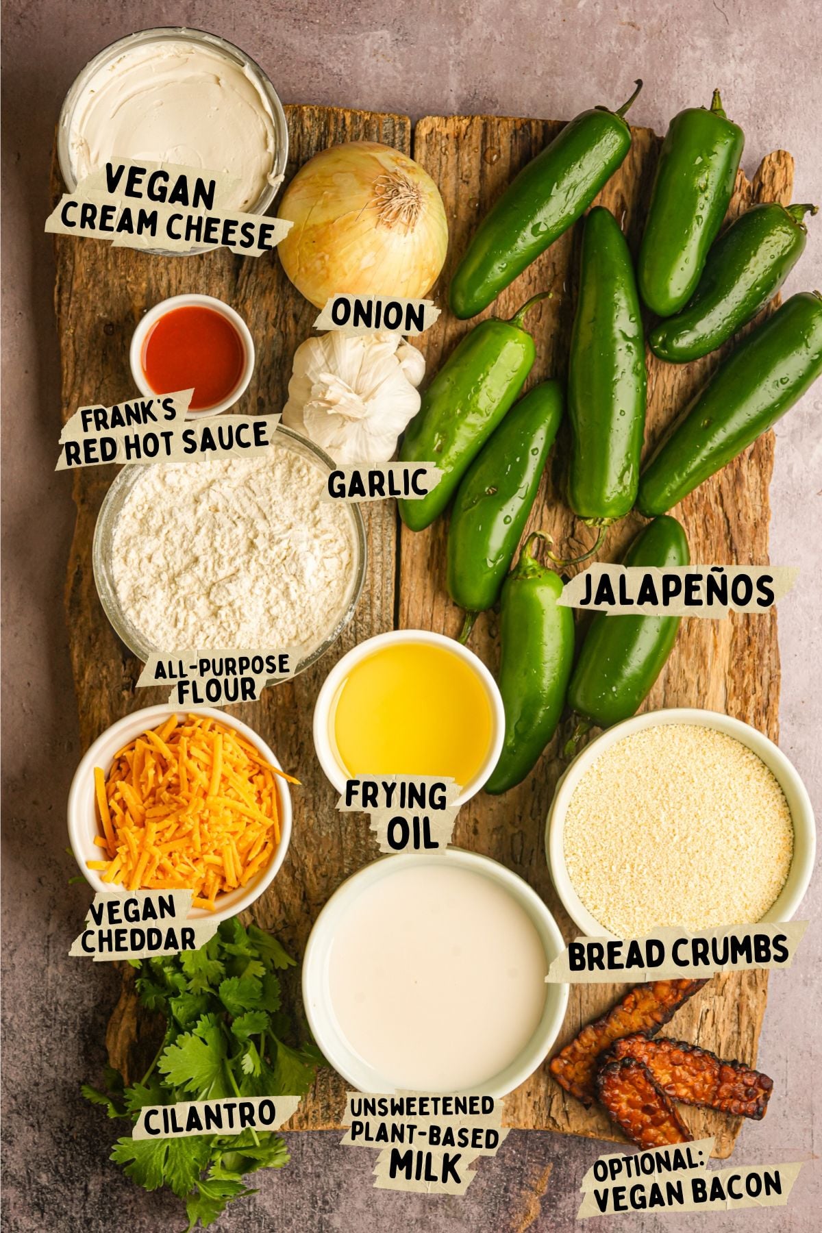 A wooden board with labeled ingredients for a vegan jalapeno popper recipe, including vegan cream cheese, hot sauce, onion, garlic, jalapenos, flour, vegan cheddar, frying oil, bread crumbs, cilantro, plant-based milk, and vegan bacon.