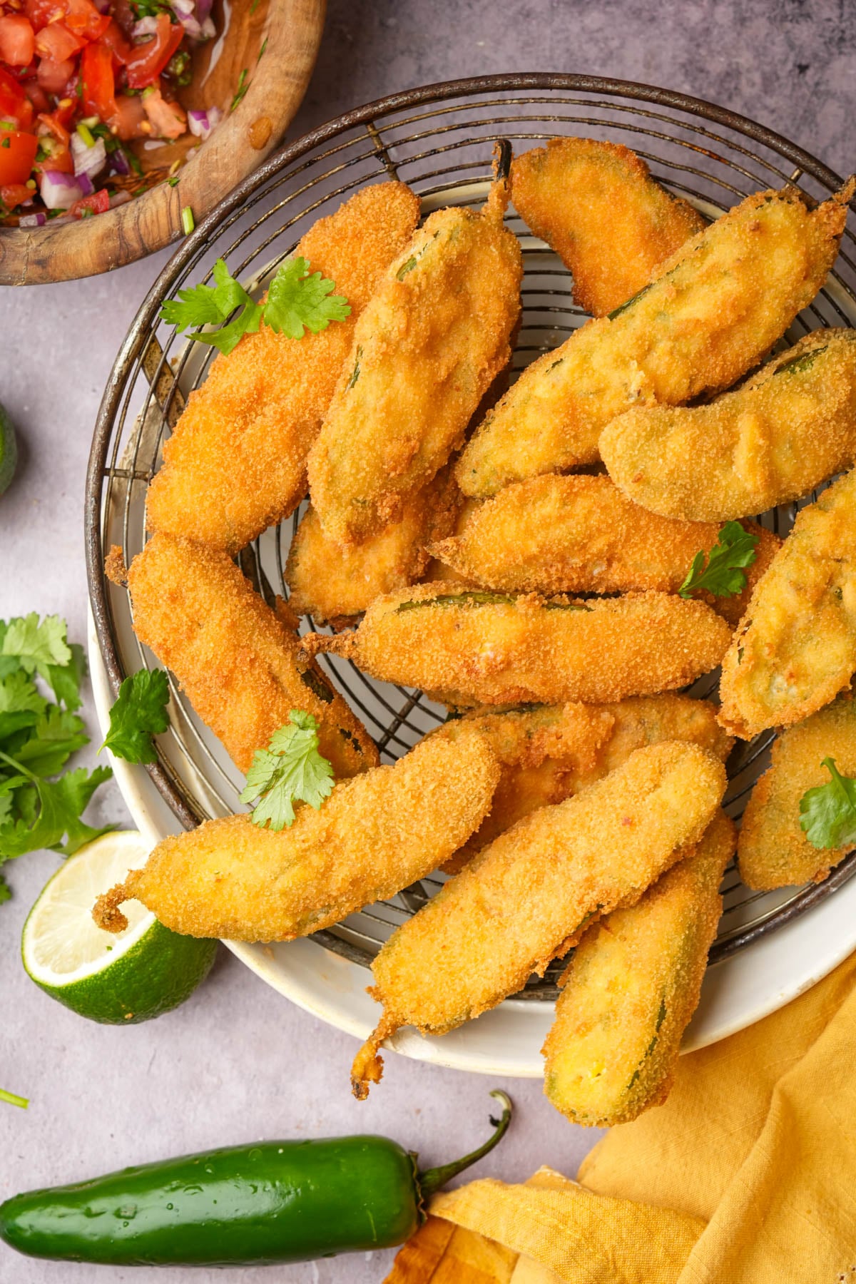A plate of golden-brown, breaded and fried vegan jalapeño poppers is arranged on a wire rack, garnished with cilantro, with a lime, a fresh jalapeño, and a bowl of salsa nearby.
