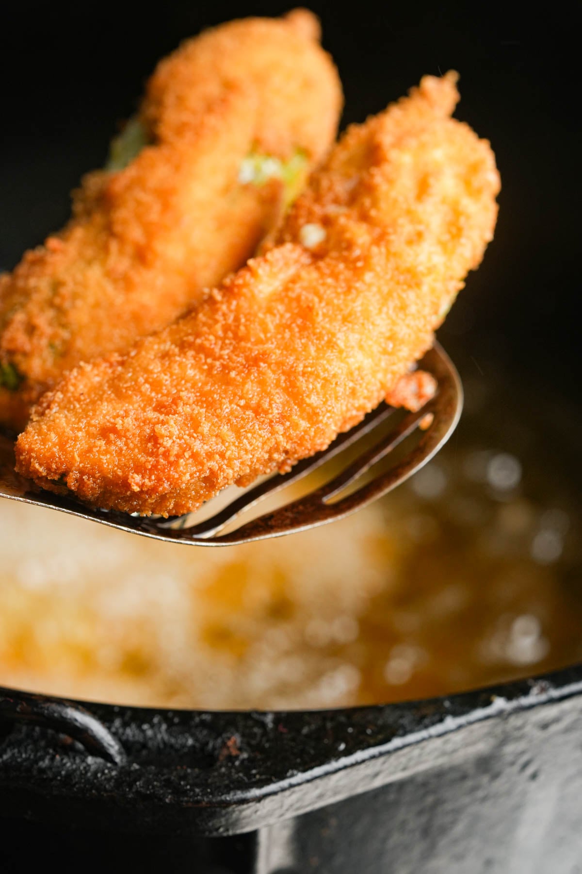 Two golden-brown breaded vegan jalapeño poppers on a slotted spoon held above bubbling hot oil in a dark pot, freshly fried and crispy.