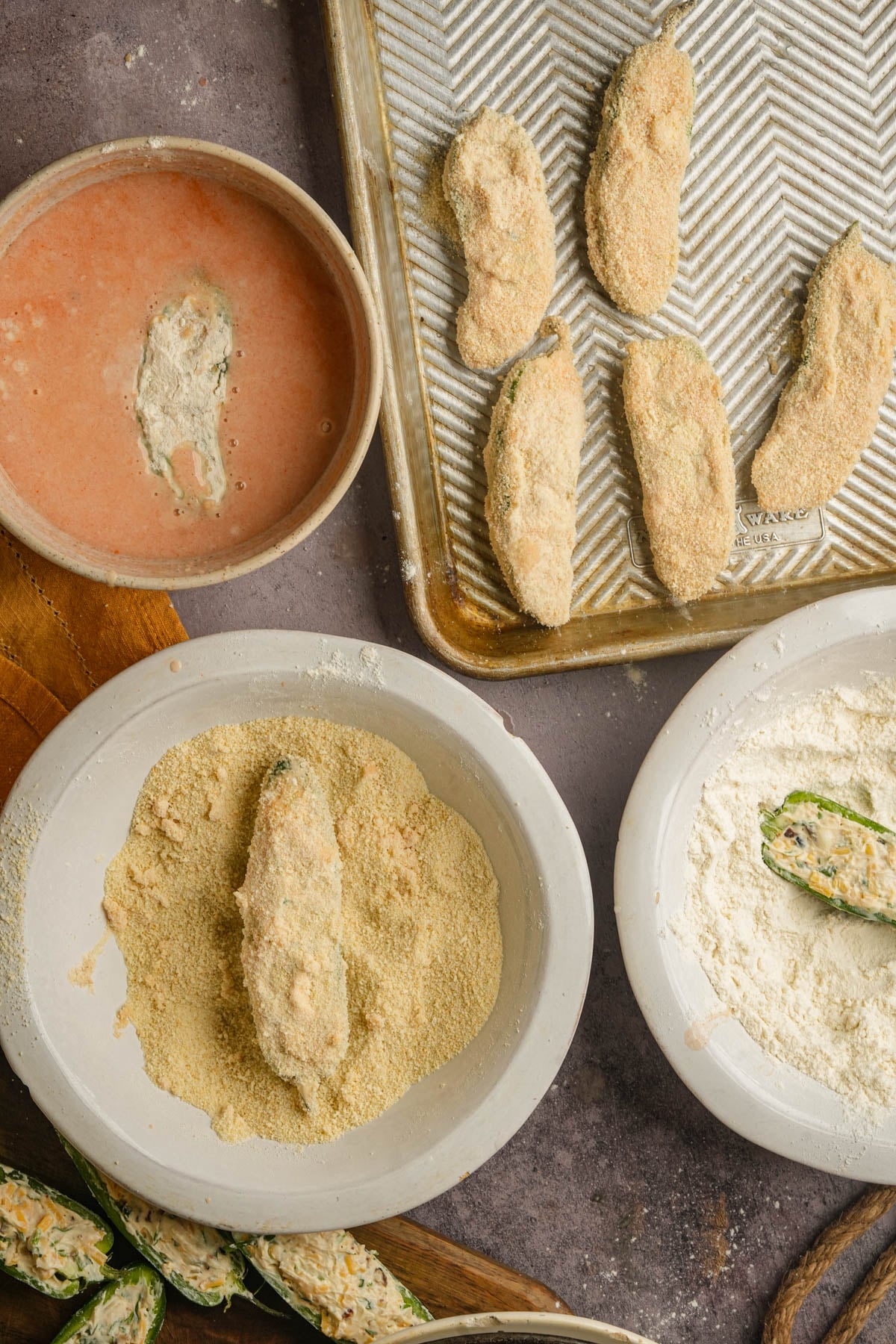 Overhead view of vegan jalapeño poppers being breaded. Bowls of flour, hot sauce/milk wash, and breadcrumbs surround a baking sheet with breaded poppers, and some halved, filled jalapeños are visible in the corner.