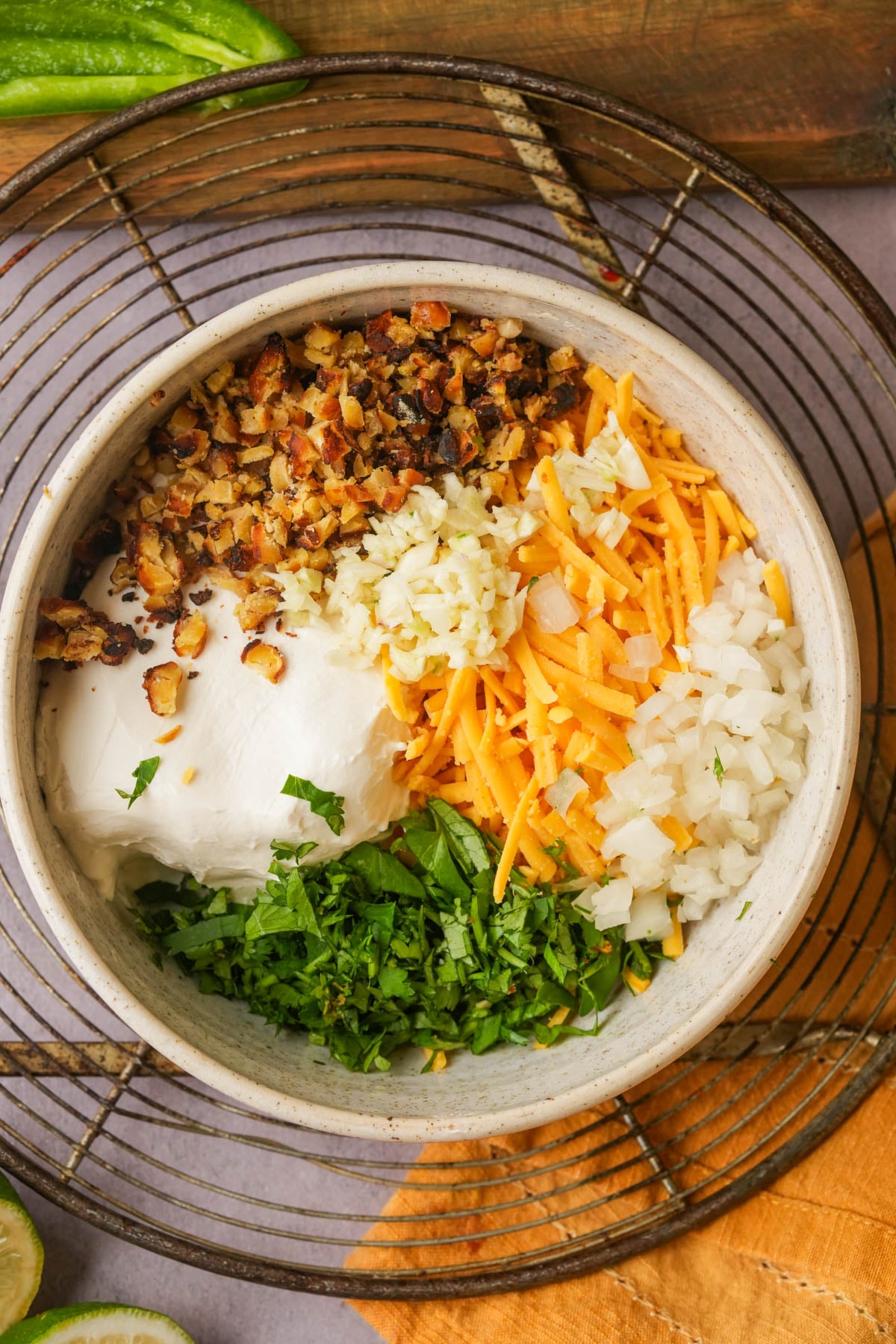 A bowl containing chopped vegan bacon, shredded cheddar cheese, minced garlic, diced onions, cream cheese, and chopped fresh cilantro, arranged in separate sections before mixing. A wire rack and napkin are underneath the bowl.