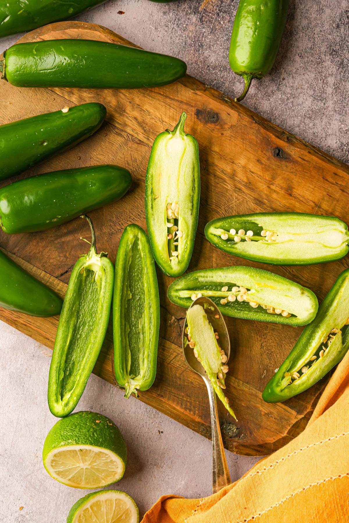 A wooden board with whole and halved fresh jalapeños, a spoon scooping seeds from one half, a sliced lime, and a yellow cloth on a gray surface.