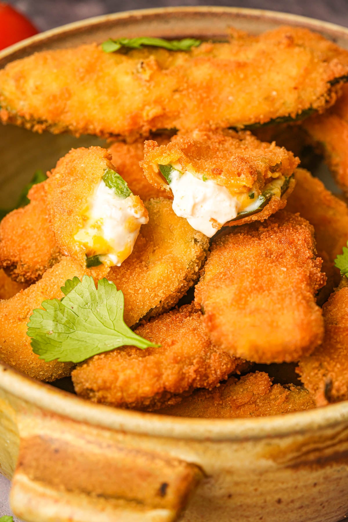 A bowl filled with golden-brown, crispy breaded vegan jalapeño poppers, some topped with cilantro, and one showing creamy cheese filling inside.