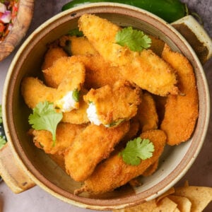 A bowl filled with golden, crispy vegan jalapeño poppers, some topped with fresh cilantro leaves, on a table with chips and colorful food nearby.
