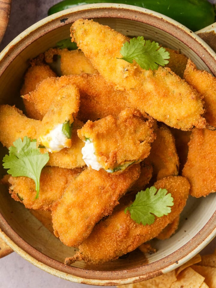 A bowl filled with golden, crispy vegan jalapeño poppers, some topped with fresh cilantro leaves, on a table with chips and colorful food nearby.