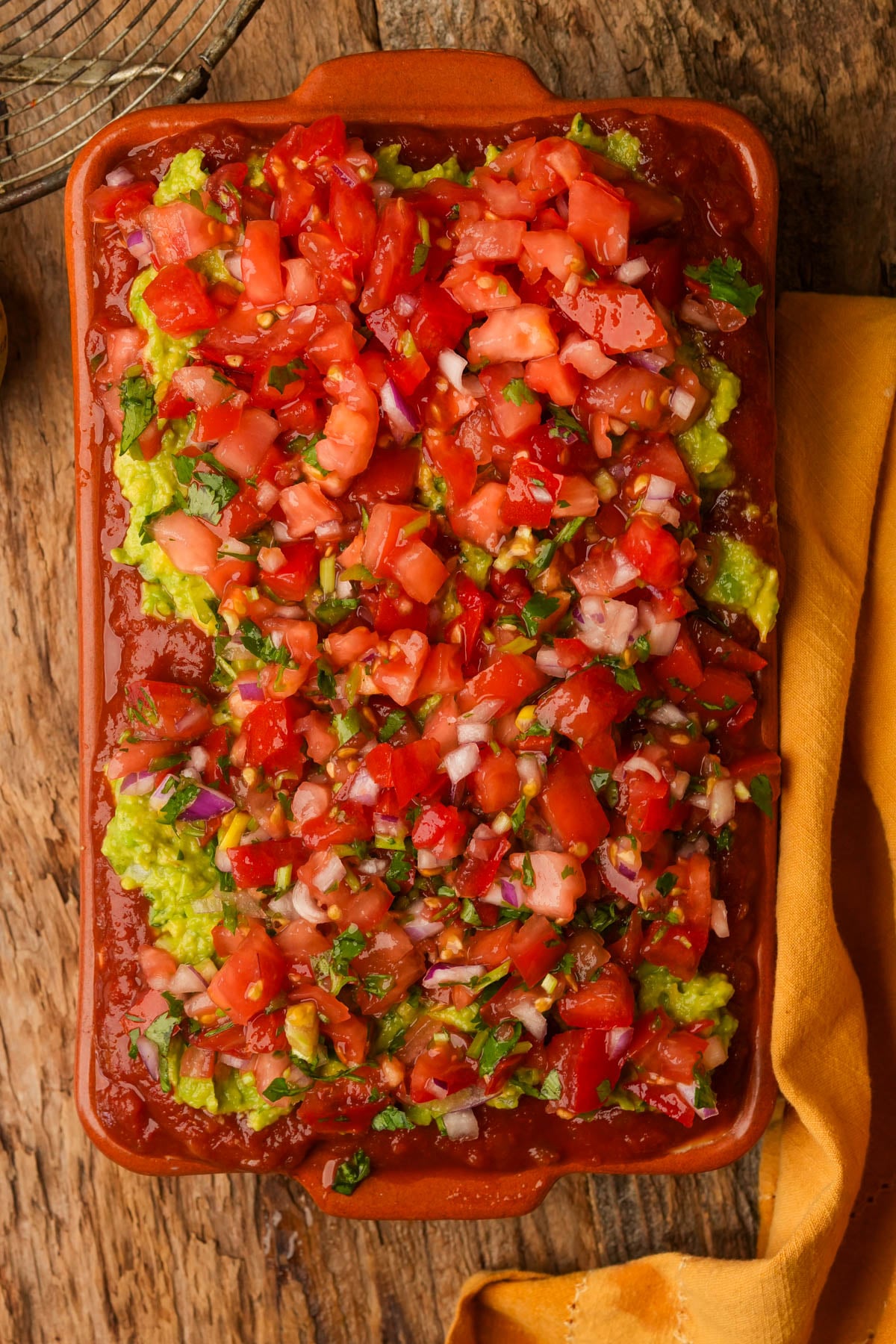 A rectangular dish holds a layered vegan seven-layer dip topped with pico de gallo.
