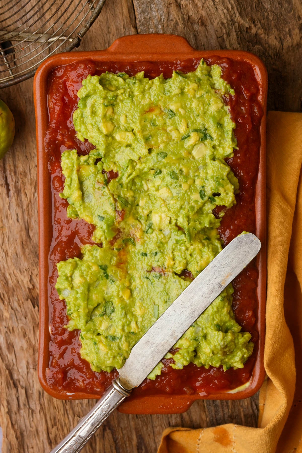 A rectangular dish filled with rvegan seven-layer dip topped with chunky guacamole sits on a wooden surface. A silver spreading knife rests inside the dish, and a yellow cloth is partially visible on the side.