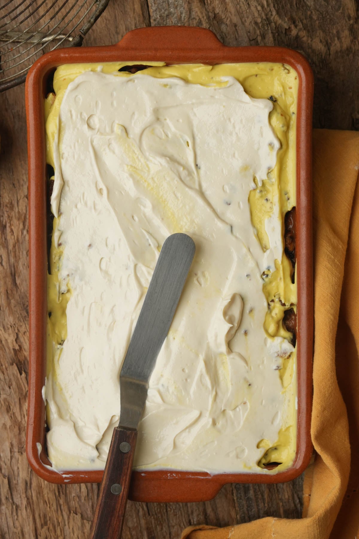 A rectangular baking dish filled with a vegan seven layer dip topped with a layer of vegan sour cream; an offset spatula rests on top. A yellow cloth is beside the dish on a wooden surface.